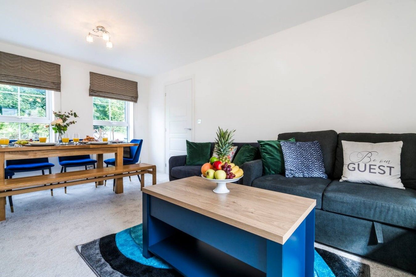 A modern living room with a dining area, a coffee table with fruit, a sofa with a 'Be our guest' pillow, and a view of greenery outside.
