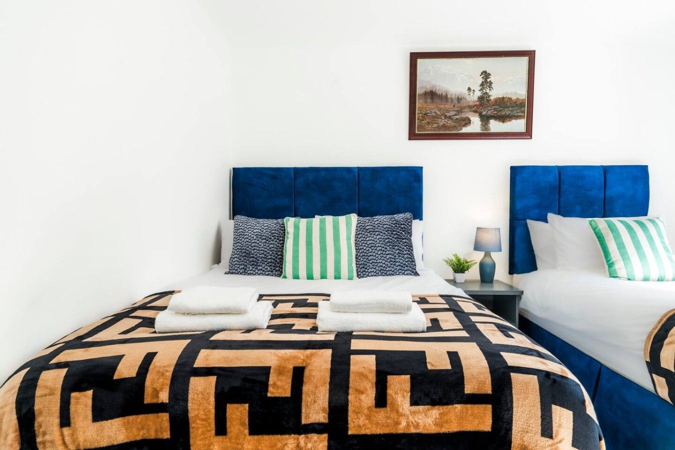 A double bed with a patterned bedspread, two pillows, a nightstand with a lamp, and a framed picture on the wall.