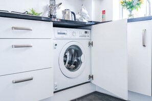 A compact, modern laundry room with a built-in washer and dryer, a countertop with kitchen utensils, and a small plant. - Argus 4-Bed Premium in Hemel: Contractor, Families (Hertfordshire)