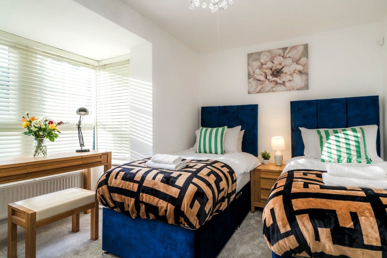 Una habitación de hotel con dos camas, un escritorio con lámpara, una banca y una ventana con persianas.
