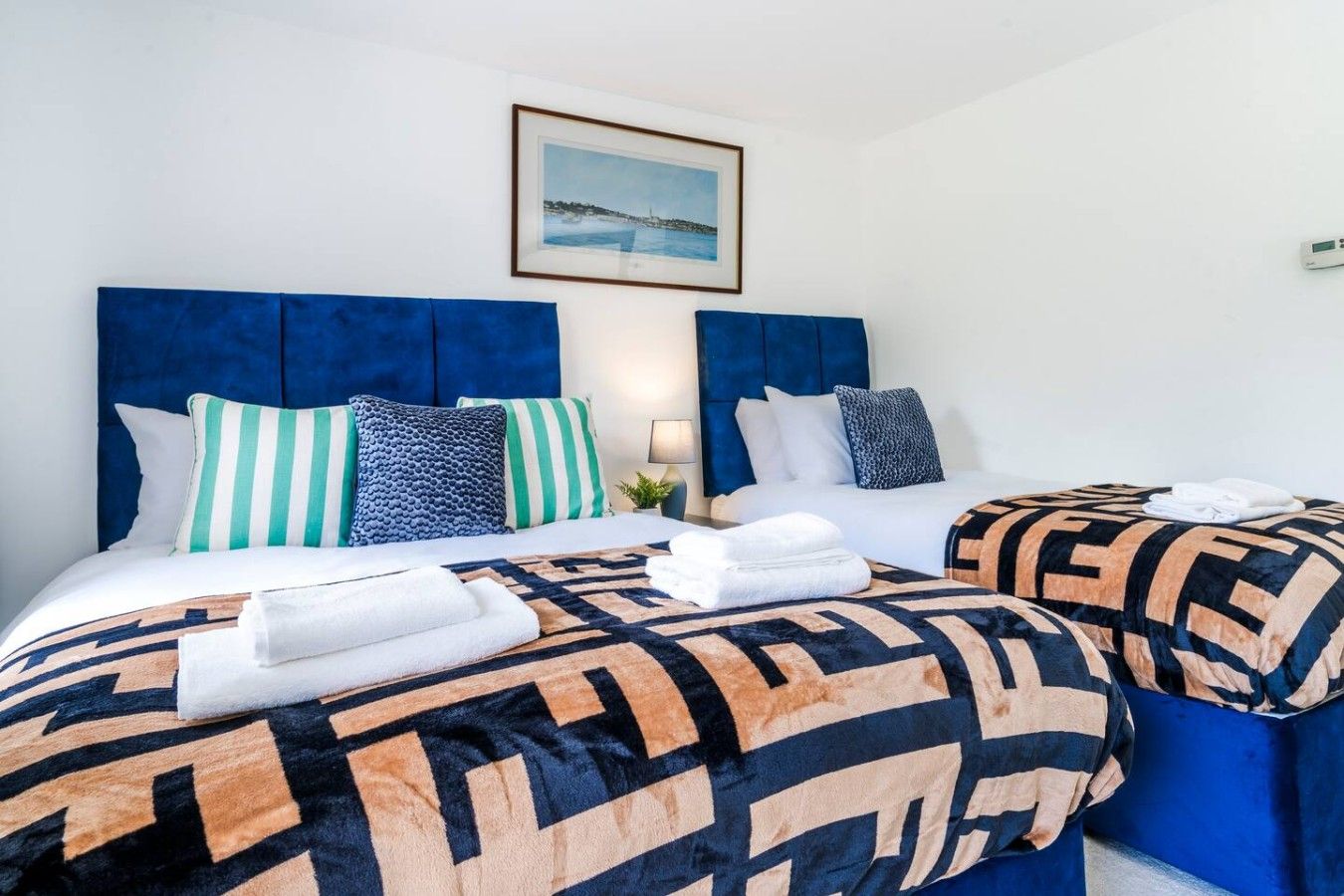 Habitación de hotel con dos camas, cada una con cabecero azul, ropa de cama blanca y almohadones estampados. Encima de las camas hay un cuadro en la pared.