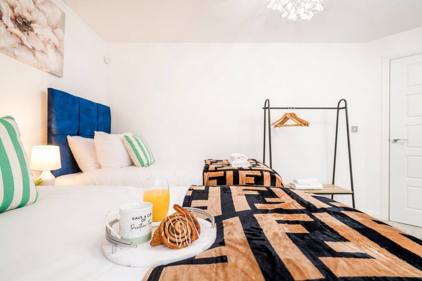 A hotel room with a bed, a hanging clothes rack, a breakfast tray with a croissant and a mug, and a wall-mounted artwork.