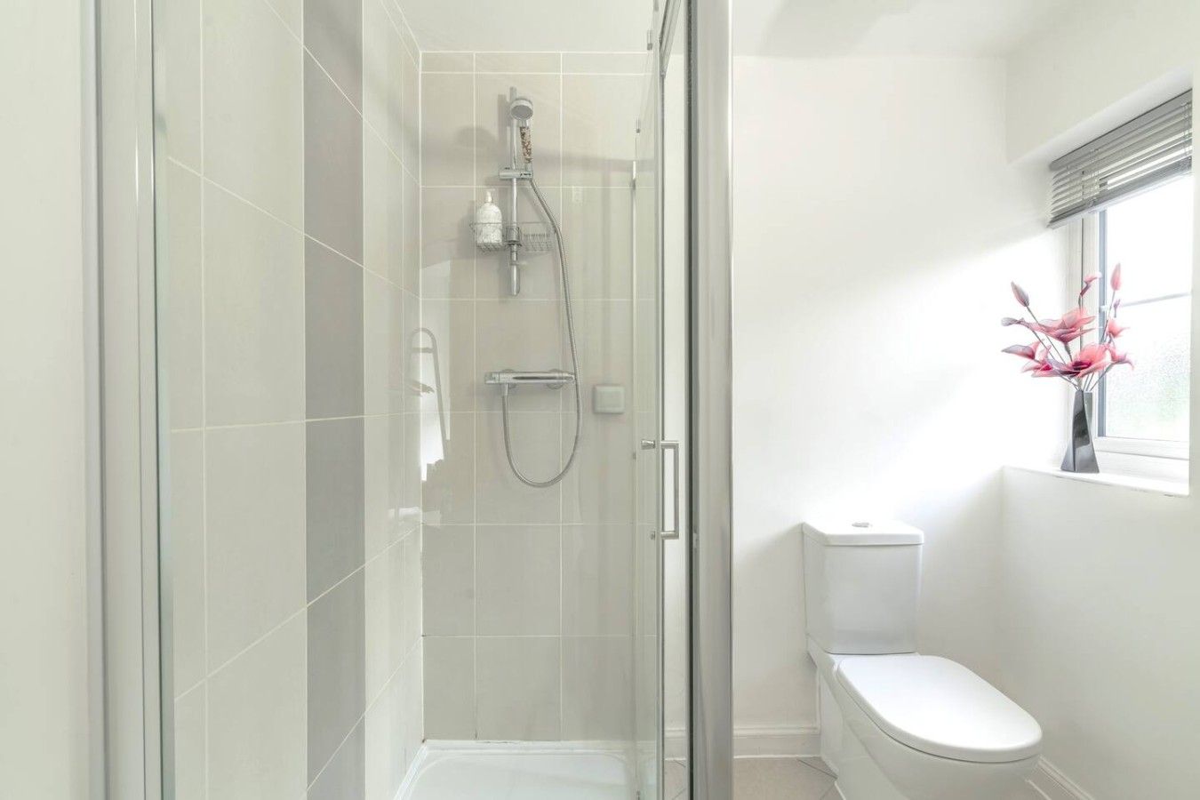 A modern bathroom with a glass shower enclosure, a toilet, and a window with a vase of flowers.