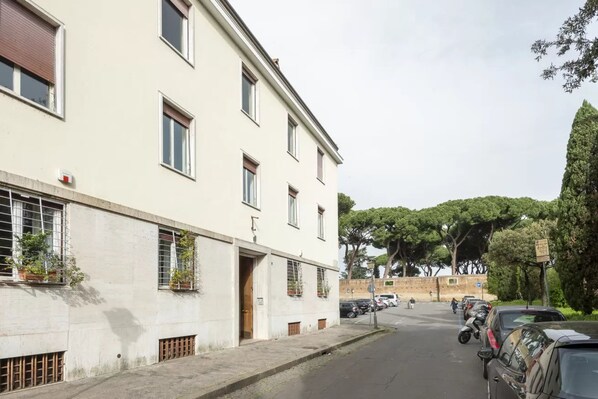 A street view with a building featuring multiple windows and a door, parked cars along the side, and trees in the background. - Aventino Luxury Apartments (Roma)