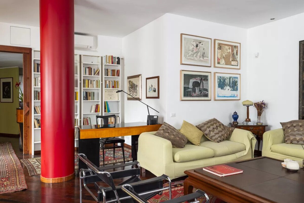 Ein Wohnzimmer mit einer roten Säule, einer Sofagarnitur, einem Couchtisch und einem Bücherregal mit Büchern.
