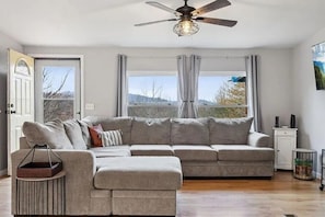 A living room with a sectional sofa, a ceiling fan, and a view through large windows. - Perfect/private location between Gatlinburg/Pigeon Forge on top of the ridge. (Sevierville)