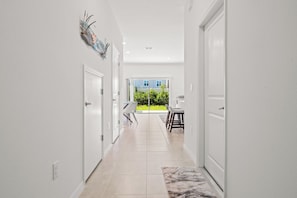 A hallway with white walls, a tiled floor, and a view of a dining area with chairs and a table. - Seaside Living Venice-Nokomis-Sarasota | 7 MIN TO THE BEACHES | GATED COMMUNITY (Nokomis)