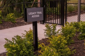 Placa indicando 'Legacy Trail Access', com um portão de metal e vegetação ao primeiro plano.