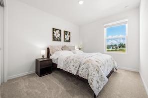 A bedroom with a bed, nightstand, lamp, two framed floral paintings, and a window with a view of palm trees. - Seaside Living Venice-Nokomis-Sarasota | 7 MIN TO THE BEACHES | GATED COMMUNITY (Nokomis)