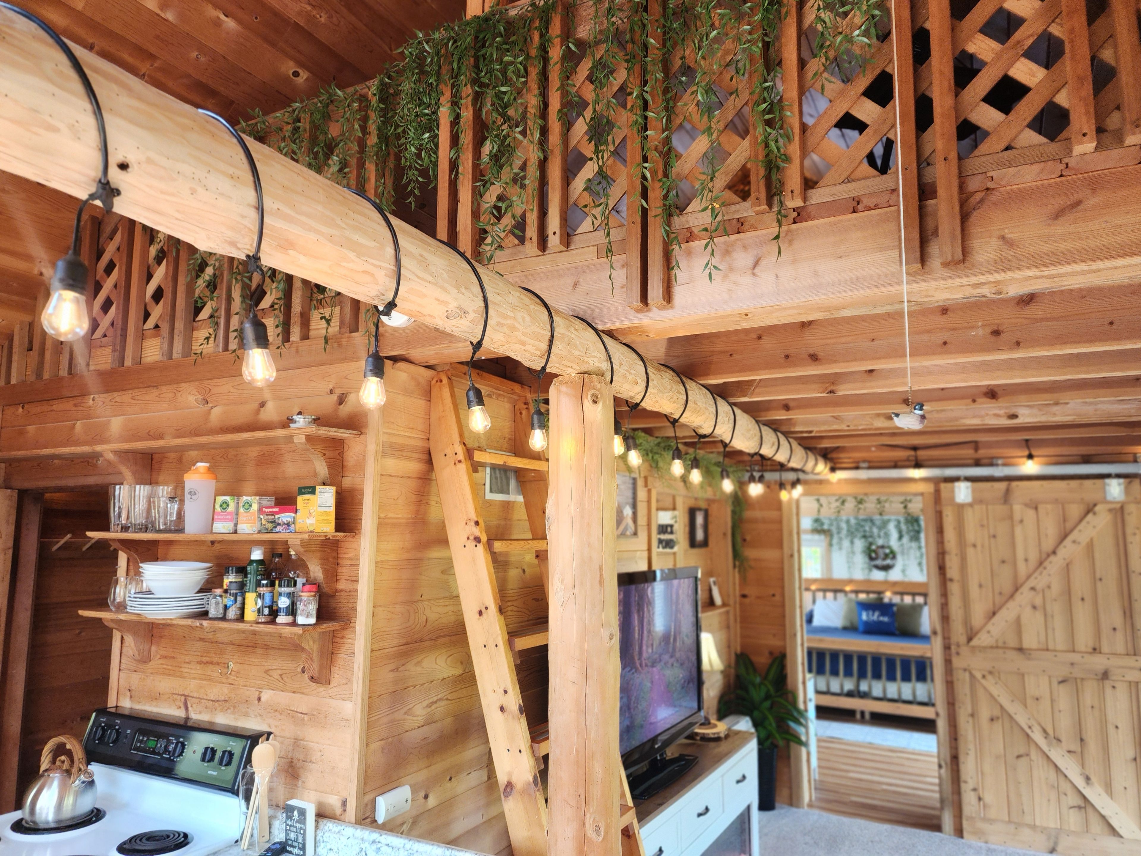 A wooden interior with a kitchen area, wooden shelves with items, and hanging lights.