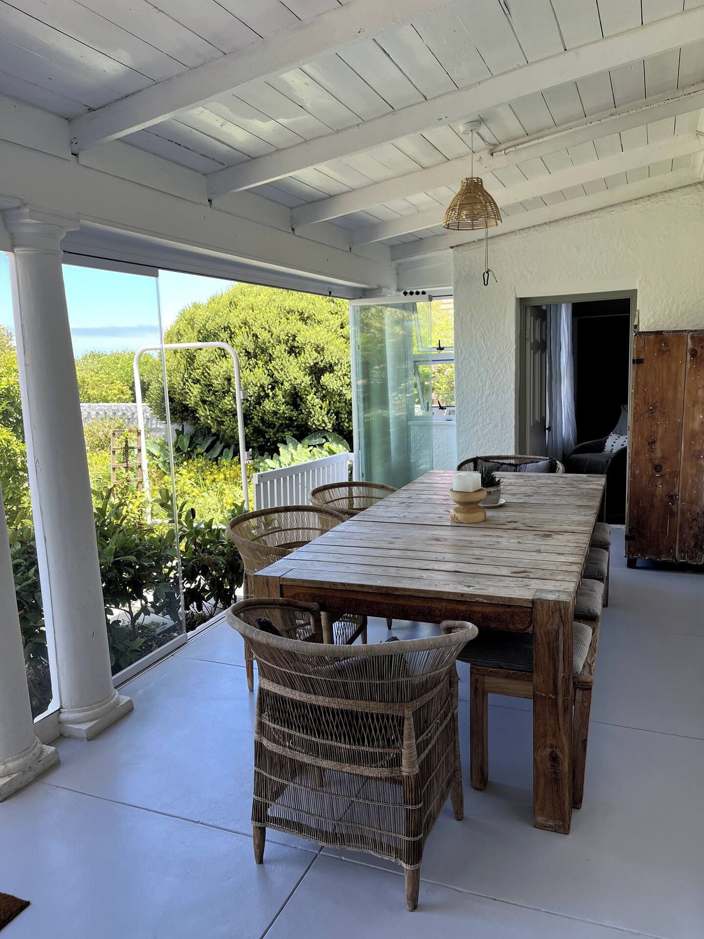 Un patio techado con una mesa de madera y sillas de mimbre, con vista a un jardín exuberante y una masa de agua.