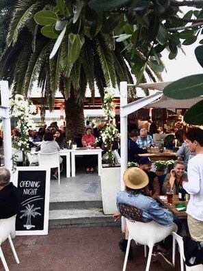 An outdoor dining area with white tables and chairs, surrounded by greenery and palm trees. - The Original Kalk Bay Farmhouse at the Homestead (Cape Town)