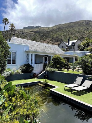 A white house with a sloped roof, a small pond, and a patio with chairs. - The Original Kalk Bay Farmhouse at the Homestead (Cape Town)
