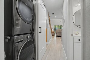 A laundry room with stacked washing machines, a hallway leading to a living area with a sofa and a view of greenery outside. - Del Lago - 3 Minutes walk to the Beach (St. Augustine)