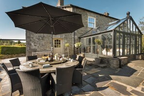 A stone house with a conservatory, outdoor dining area with wicker furniture, and a large umbrella. - Three Mile House (Carnforth)