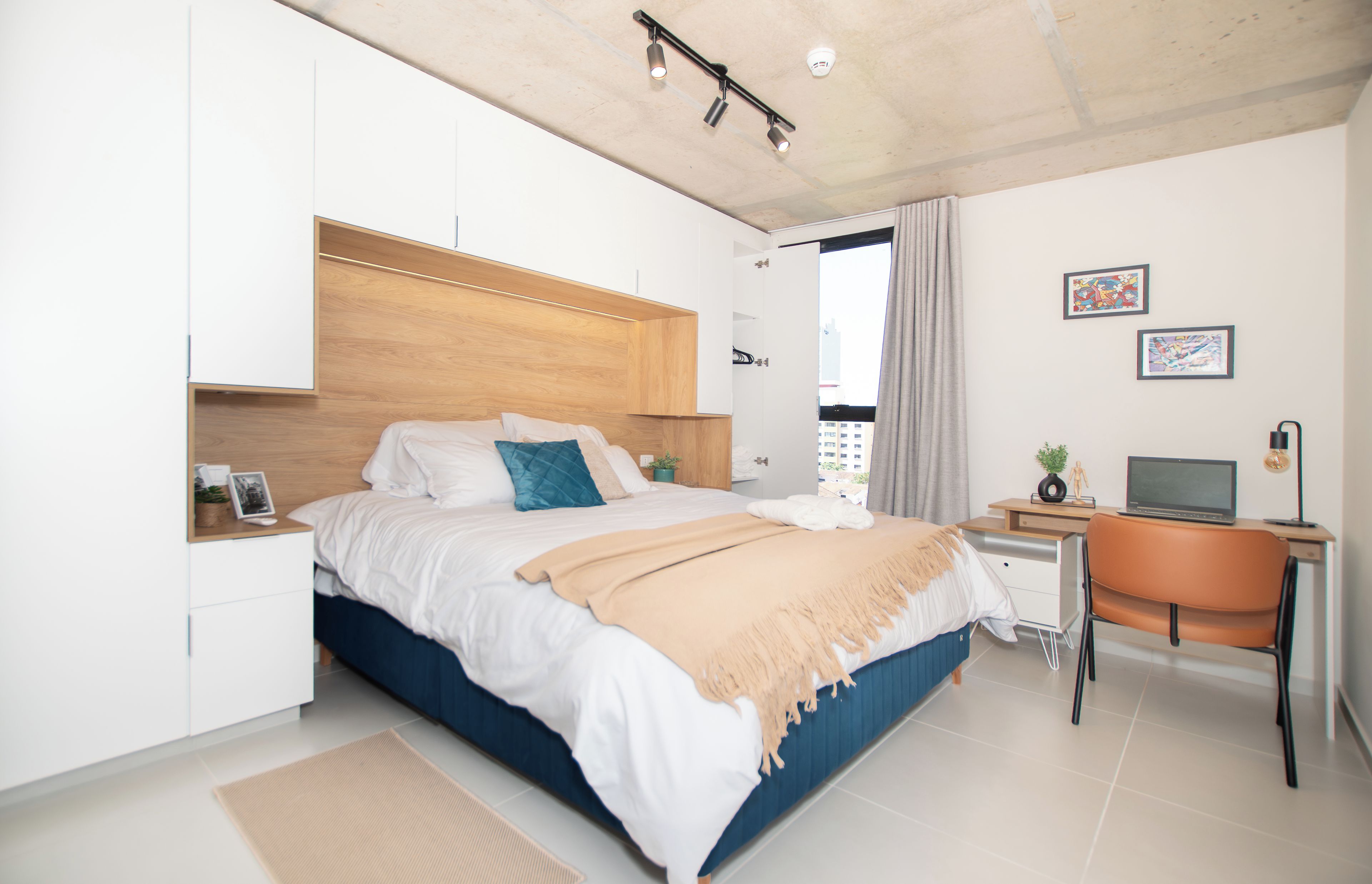 Un dormitorio moderno con cama, escritorio y silla, que ofrece vistas a la ciudad a través del ventanal.