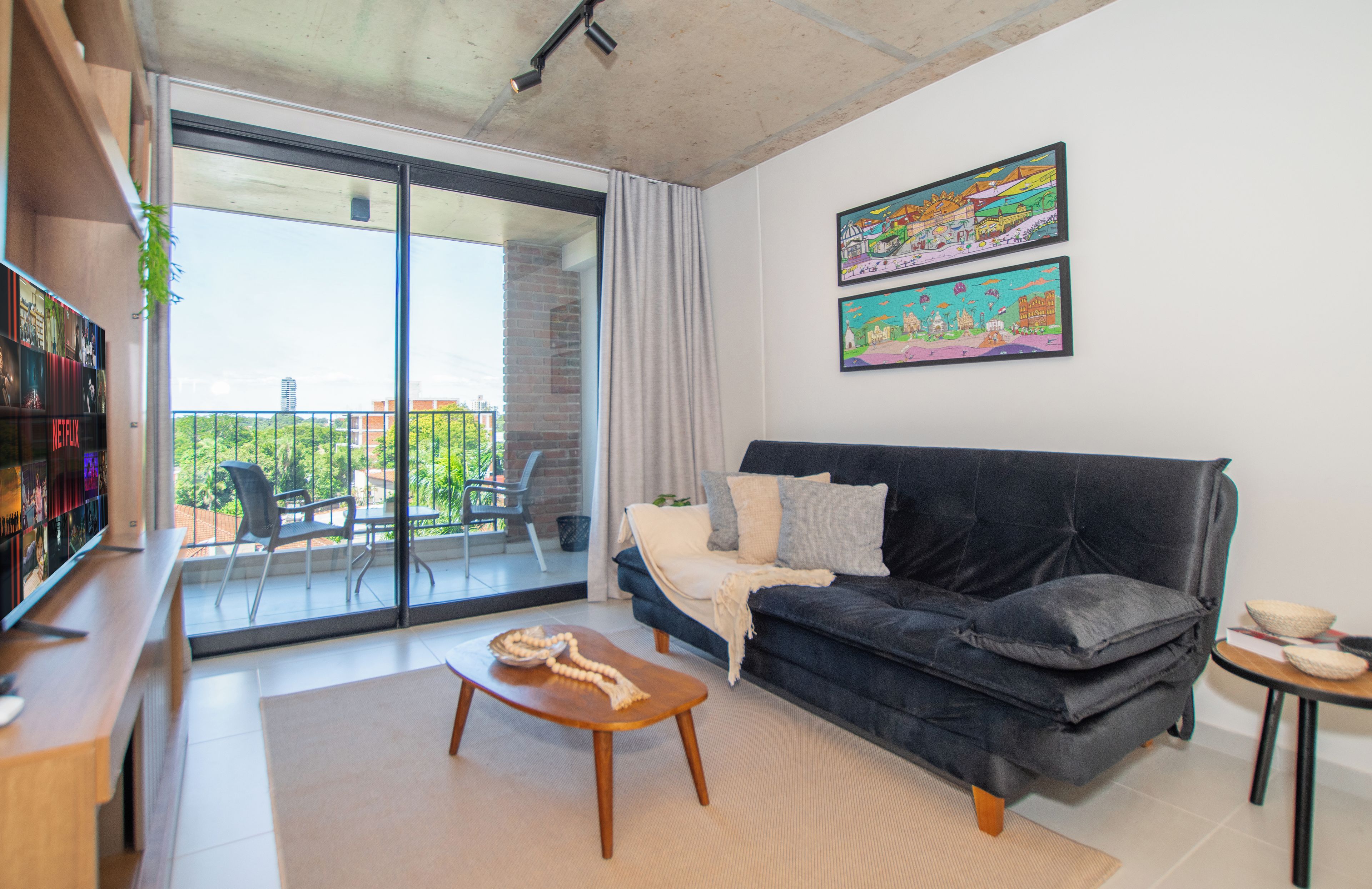 Una sala de estar moderna con un sofá, una mesa de centro y un televisor sobre un mueble bajo. Hay un balcón con vistas a zonas verdes y edificios.
