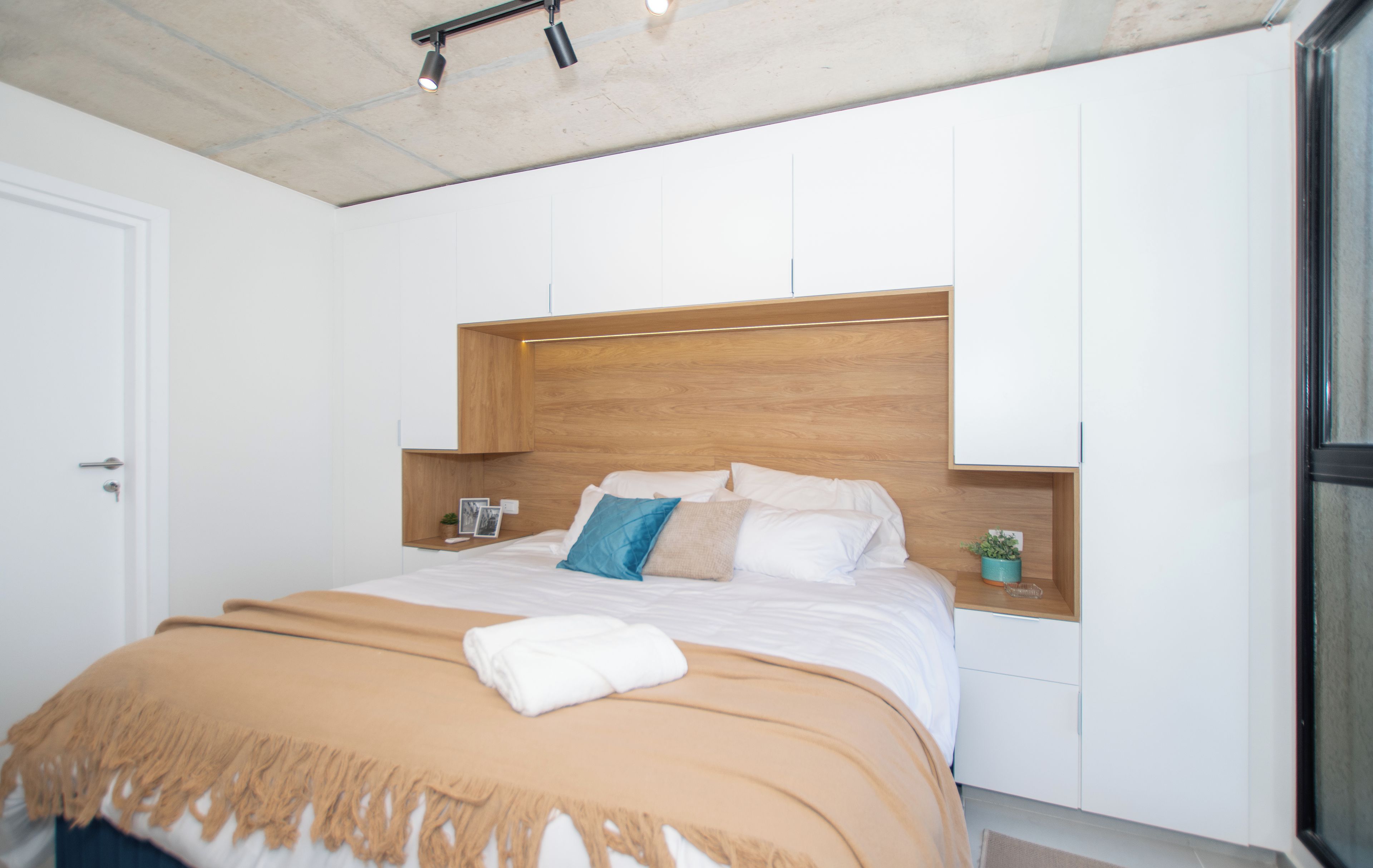 A modern bedroom with a wooden headboard, a bed with white bedding, a beige throw blanket, and a small bedside table with a plant.