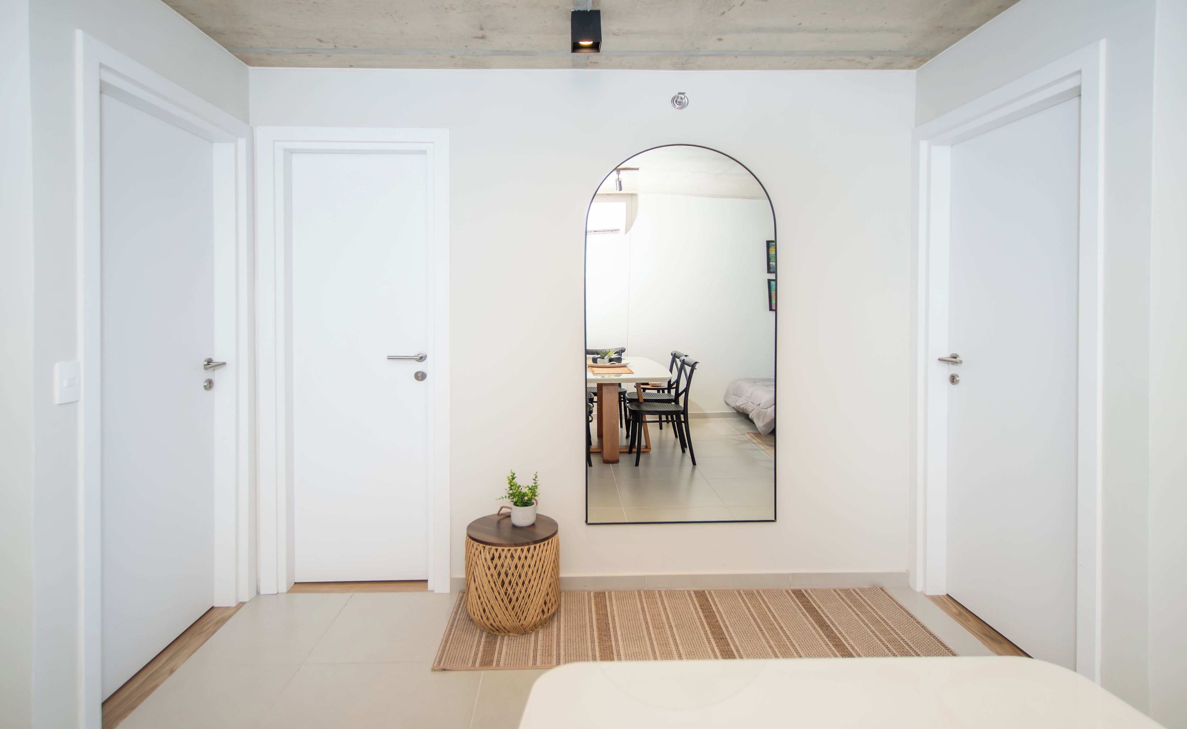 A bedroom with a large mirror, a bed, a small table with a plant, and a view of a dining area through the mirror.