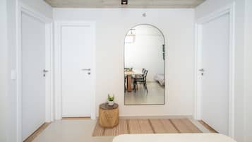 A bedroom with a large mirror, a bed, a small table with a plant, and a view of a dining area through the mirror.