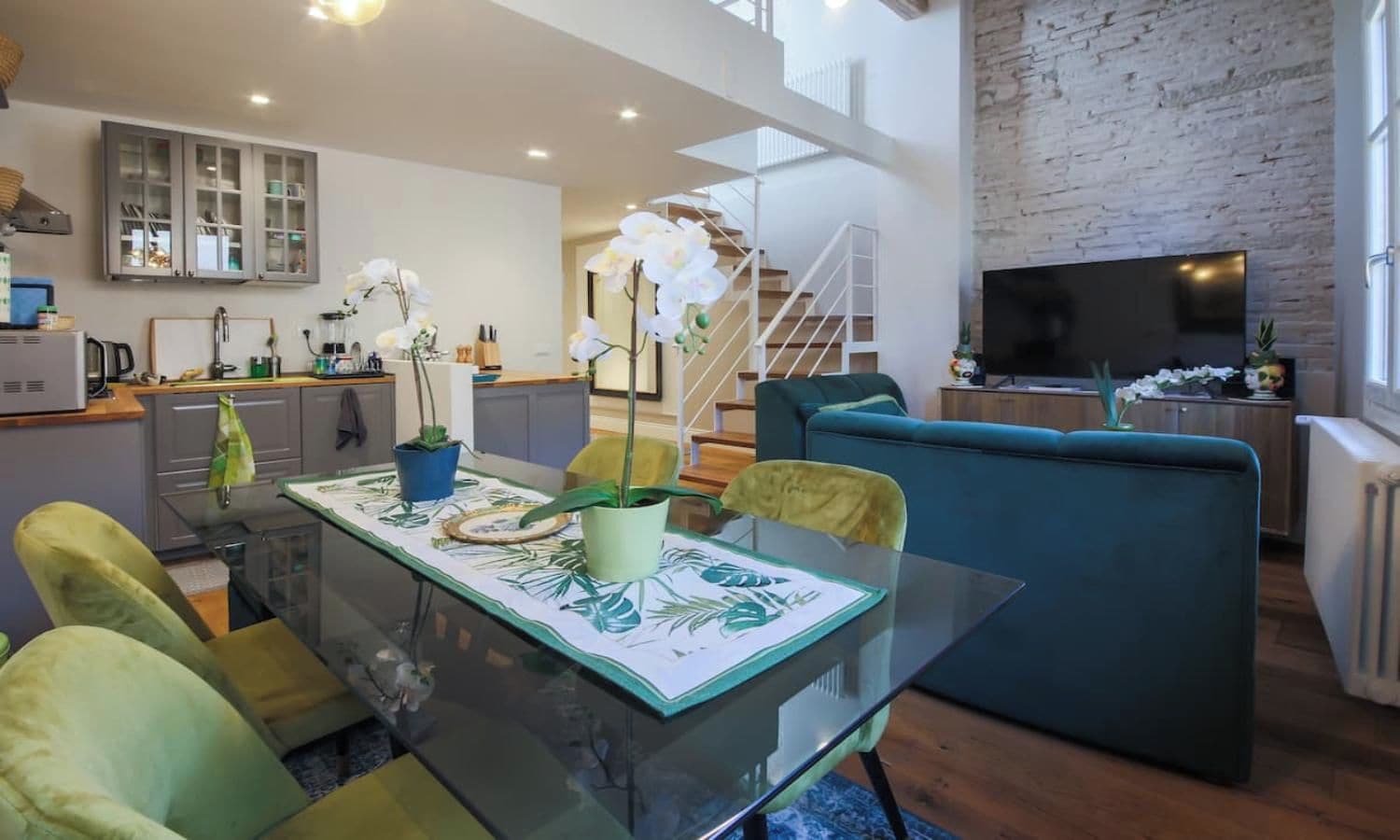 A modern living space with a glass dining table, green chairs, a blue sofa, and a kitchen area in the background.