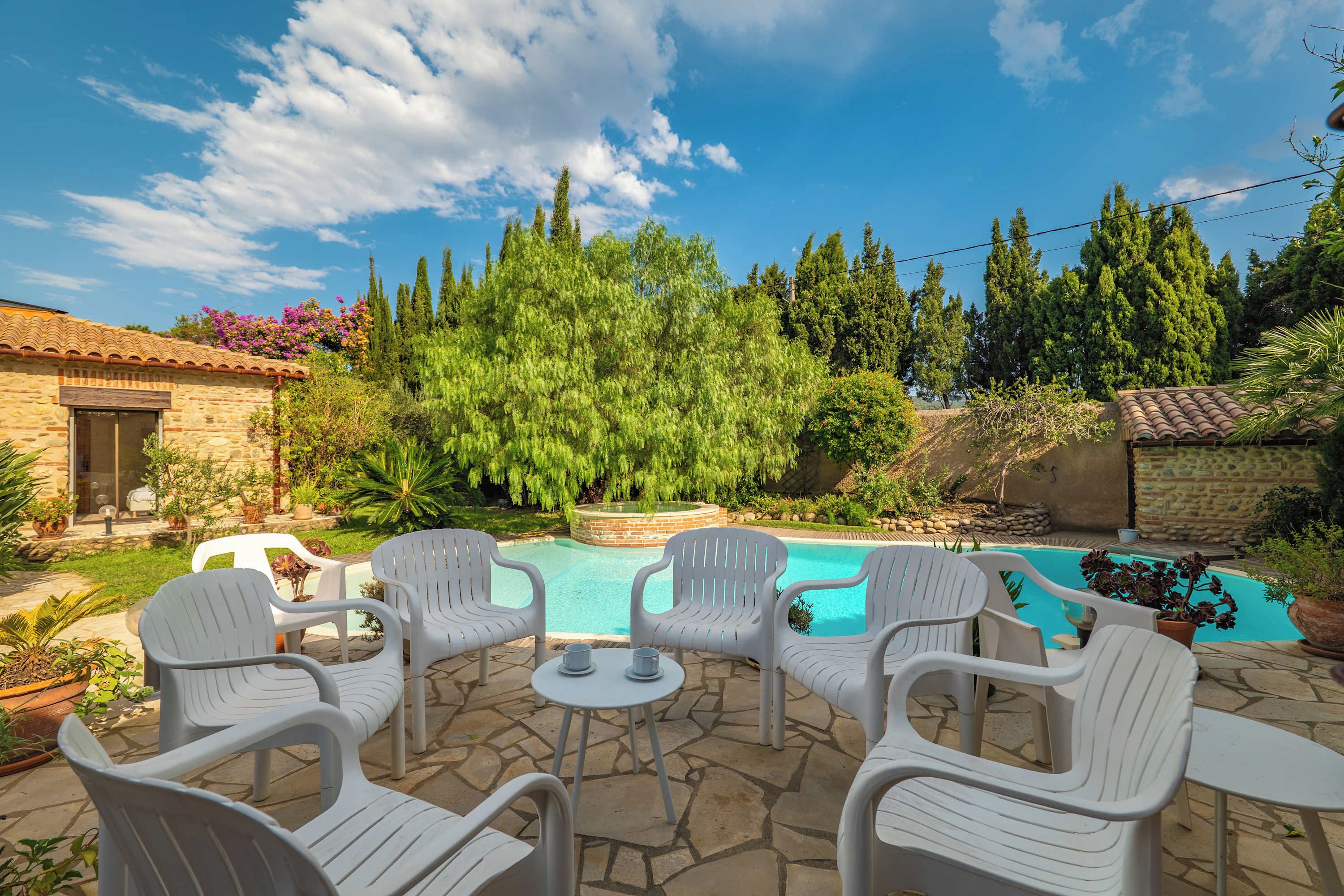 A backyard with a swimming pool, surrounded by stone patio, white plastic chairs, and lush greenery.