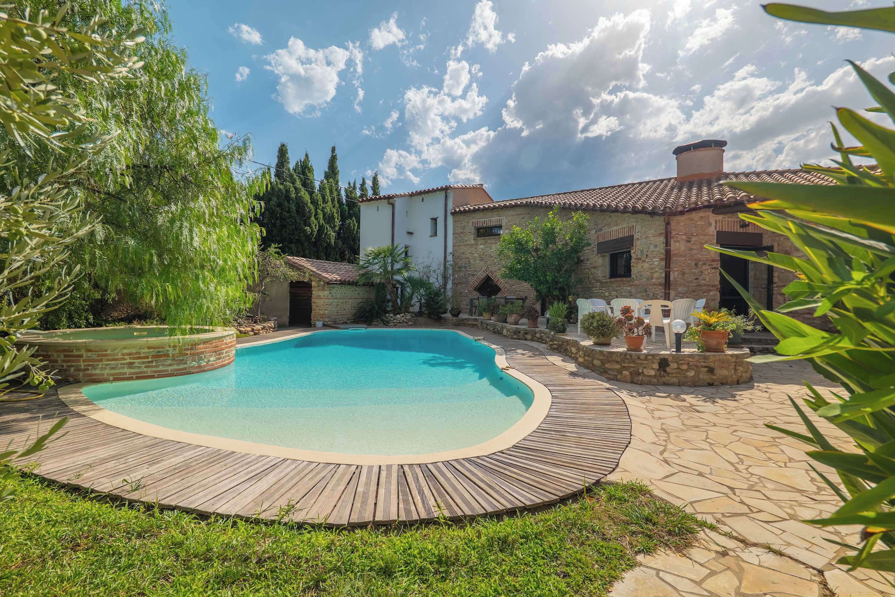 A swimming pool with a wooden deck surrounding it, set against a backdrop of stone buildings and greenery.