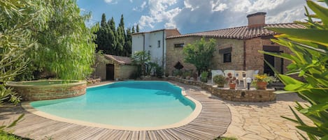 A swimming pool with a wooden deck surrounding it, set against a backdrop of stone buildings and greenery.