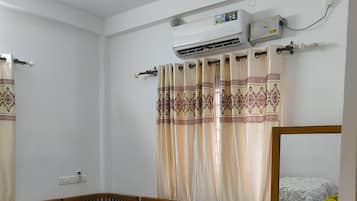 Una habitación con un sofá de madera, una mesita pequeña, un mueble de madera con cajones, un aire acondicionado de pared y una cortina con un borde estampado.