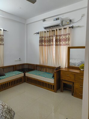 Ein Zimmer mit einer Holzsofa, einem kleinen Tisch, einer Kommode mit Schubladen, einer Wandklimaanlage und einem Vorhang mit gemustertem Rand.