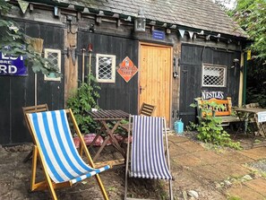 A rustic wooden building with a door, two windows, and a sign that reads 'John Bull Tyres.' There are two striped chairs and a small table outside. - Remarkable Boultons Barn With hot tub and opt Sauna (Newton Abbot)