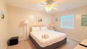 A bedroom with a bed, a ceiling fan, a nightstand with a lamp, a window with blinds, and a wall with a motivational quote. - Relaxing 2BR Condo | First Floor | Screened Porch (Myrtle Beach)