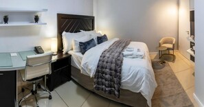 A hotel room with a bed, bedside table, lamp, desk, chair, and a small shelf with plants. - Imperial Ventures Apartments - 1 BR apt 2 (Sandton)