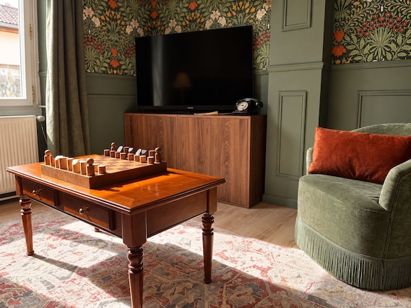 A wooden chess table in a room with a green armchair, a television, and patterned wallpaper.