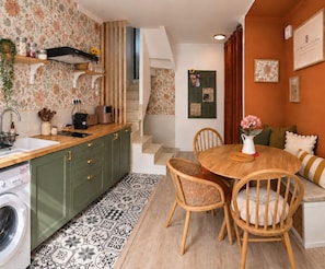 A kitchen with a sink, wooden cabinets, and a washing machine. There is a dining area with a table and chairs, and a staircase leading to another level.