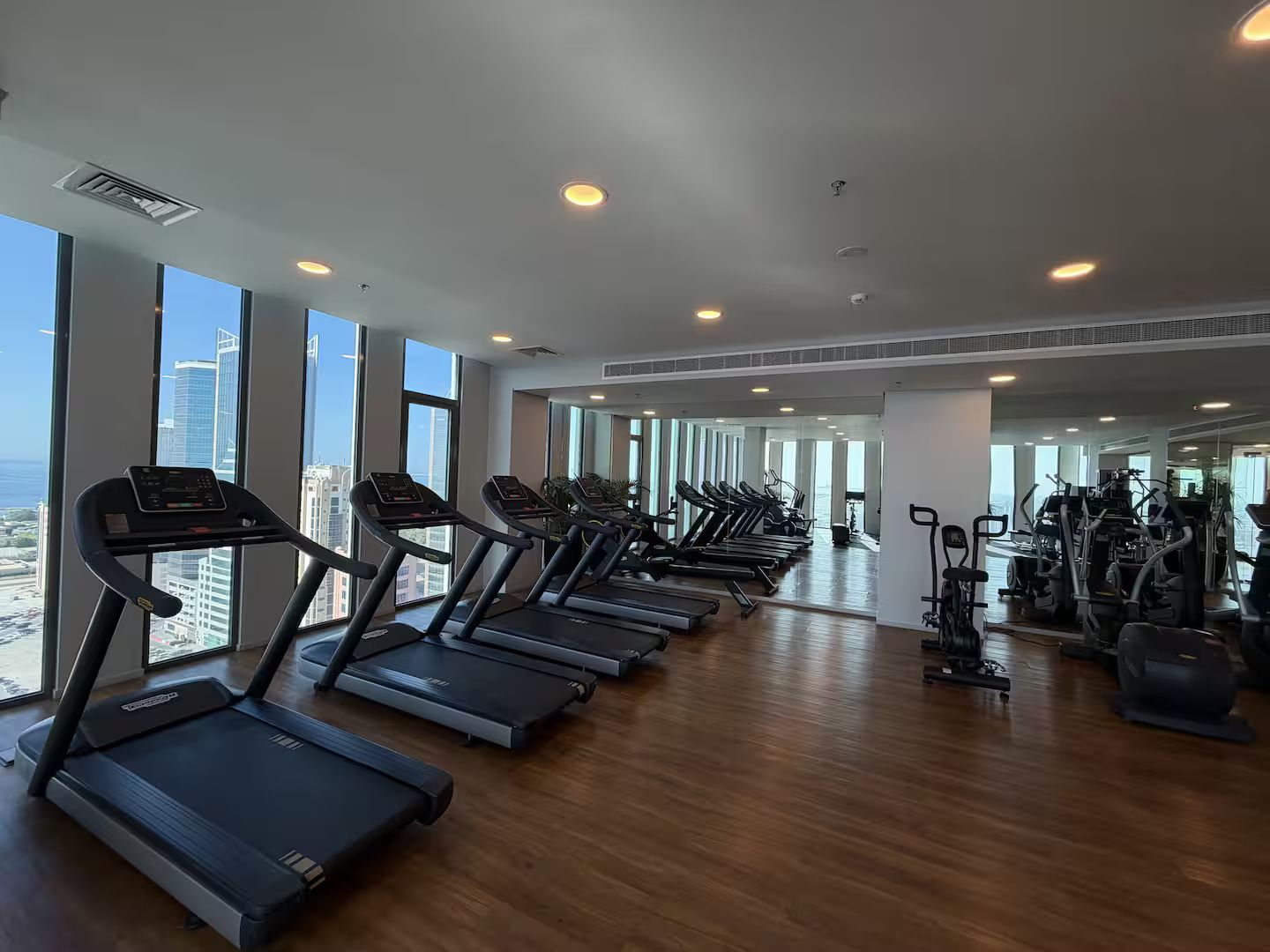 Une salle de sport avec des tapis de course alignés le long d’un mur, et de grandes fenêtres donnant sur la ville.
