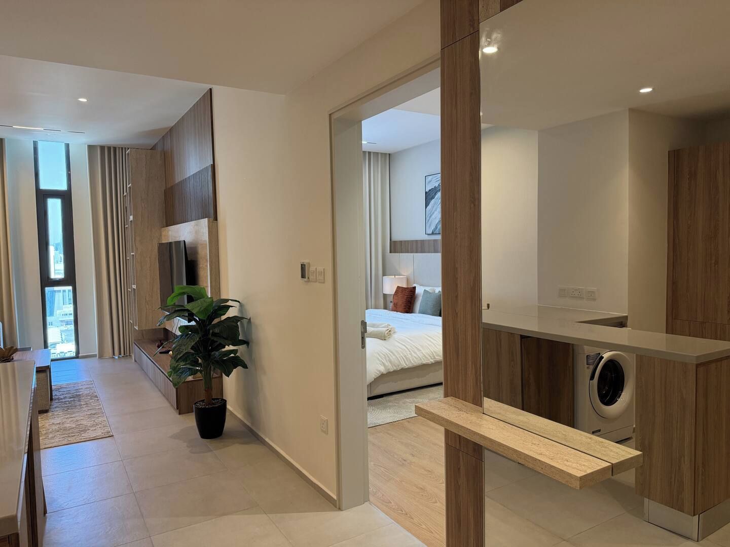 A modern hotel room with a large mirror, a washing machine, a bed with pillows, and a potted plant.