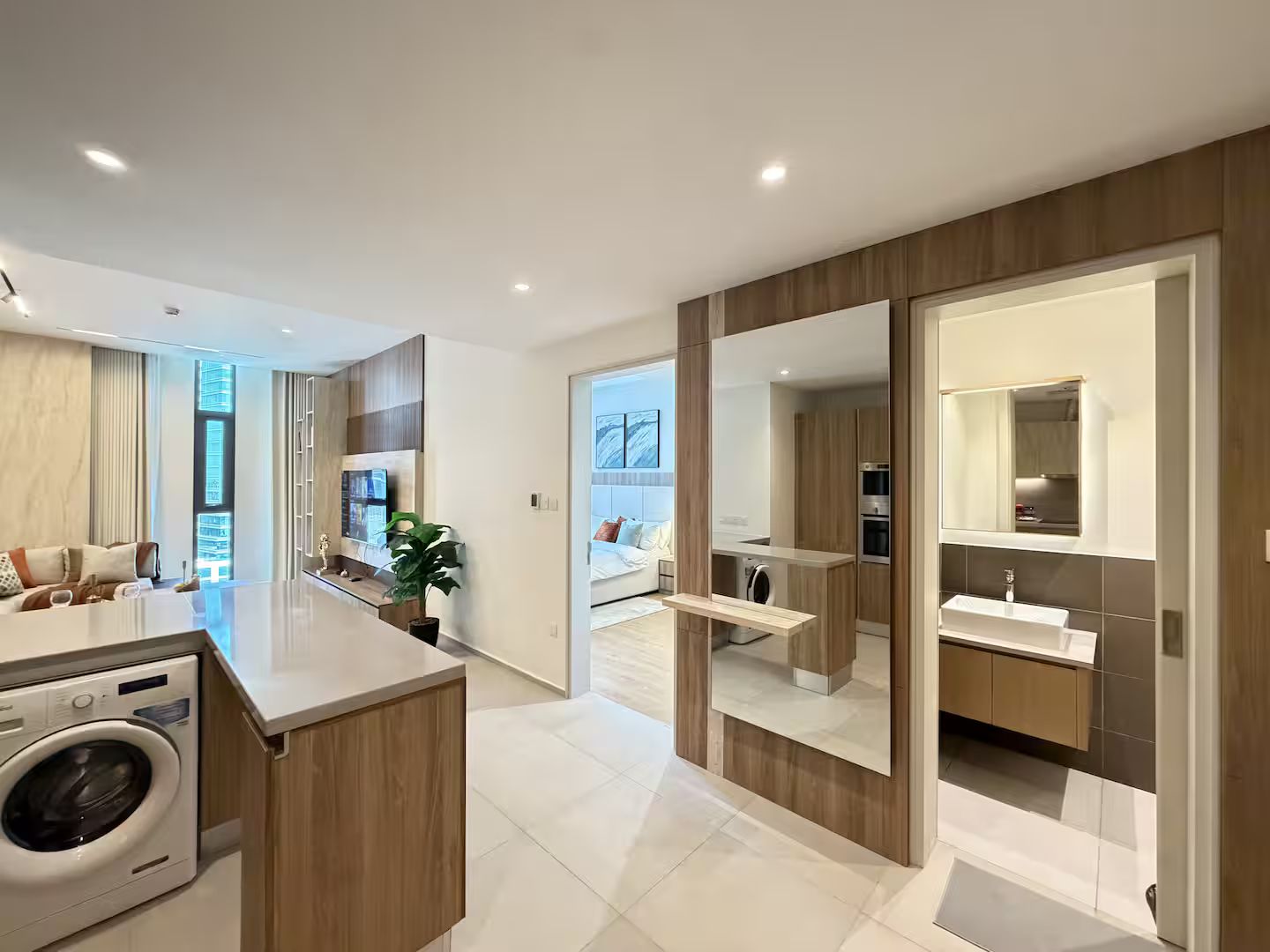 A modern apartment with a central island, a built-in washer, a large mirror, and a view into the living room.
