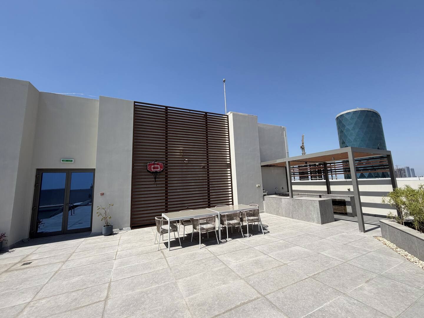 An outdoor patio area with a table and chairs, a basketball hoop, and a building with a modern design.