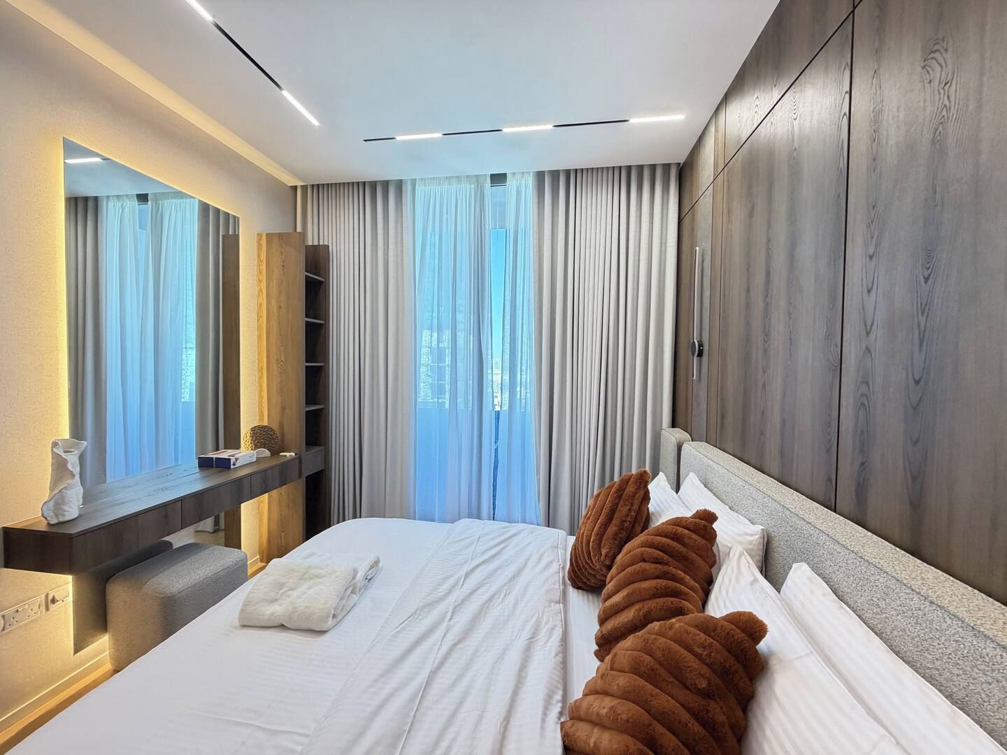 A modern hotel room with a large bed, a wooden wardrobe, a bedside table, and a large window with curtains.