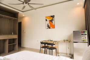 Habitació d'hotel amb un llit, una escriptori amb cadires, un ventilador de sostre, un frigorífic penjat a la paret i un quadre.
