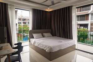 Ein Hotelzimmer mit Bett, Schreibtisch und Stuhl. Es gibt einen Balkon mit Blick auf Gebäude und Grünflächen.