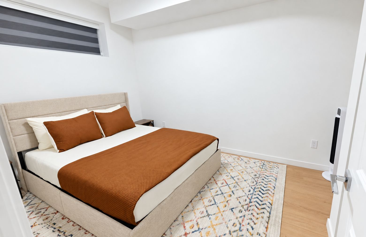 A bedroom with a bed, a nightstand, a patterned rug, and a window with blinds.
