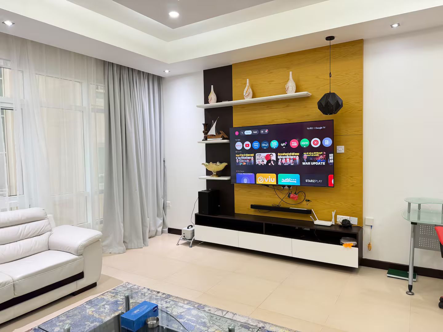 Una sala de estar moderna con un televisor de pantalla plana montado en una pared de madera decorativa, un sofá blanco y una mesa de centro de vidrio.