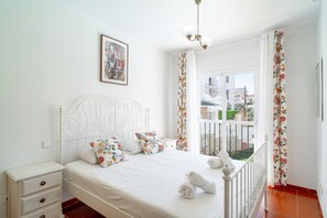 A bedroom with a white bed, floral curtains, a view of buildings, and a ceiling light. - Holiday Home in Nerja With Sea Views (Nerja)