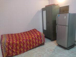 A small room with a bed covered in a floral-patterned blanket, a grey wardrobe, and a refrigerator.