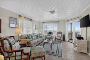 A spacious living room with a high ceiling, a television mounted on the wall, and a view of the outdoors through large windows. - Award-Winning Resort Amenities | Port O' Call A204 (Isle Of Palms)