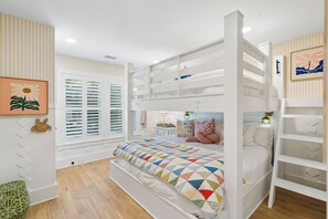 Ein Mehrbettzimmer mit farbenfroher Steppdecke, einem Fenster mit Jalousien und einem gerahmten Bild an der Wand.