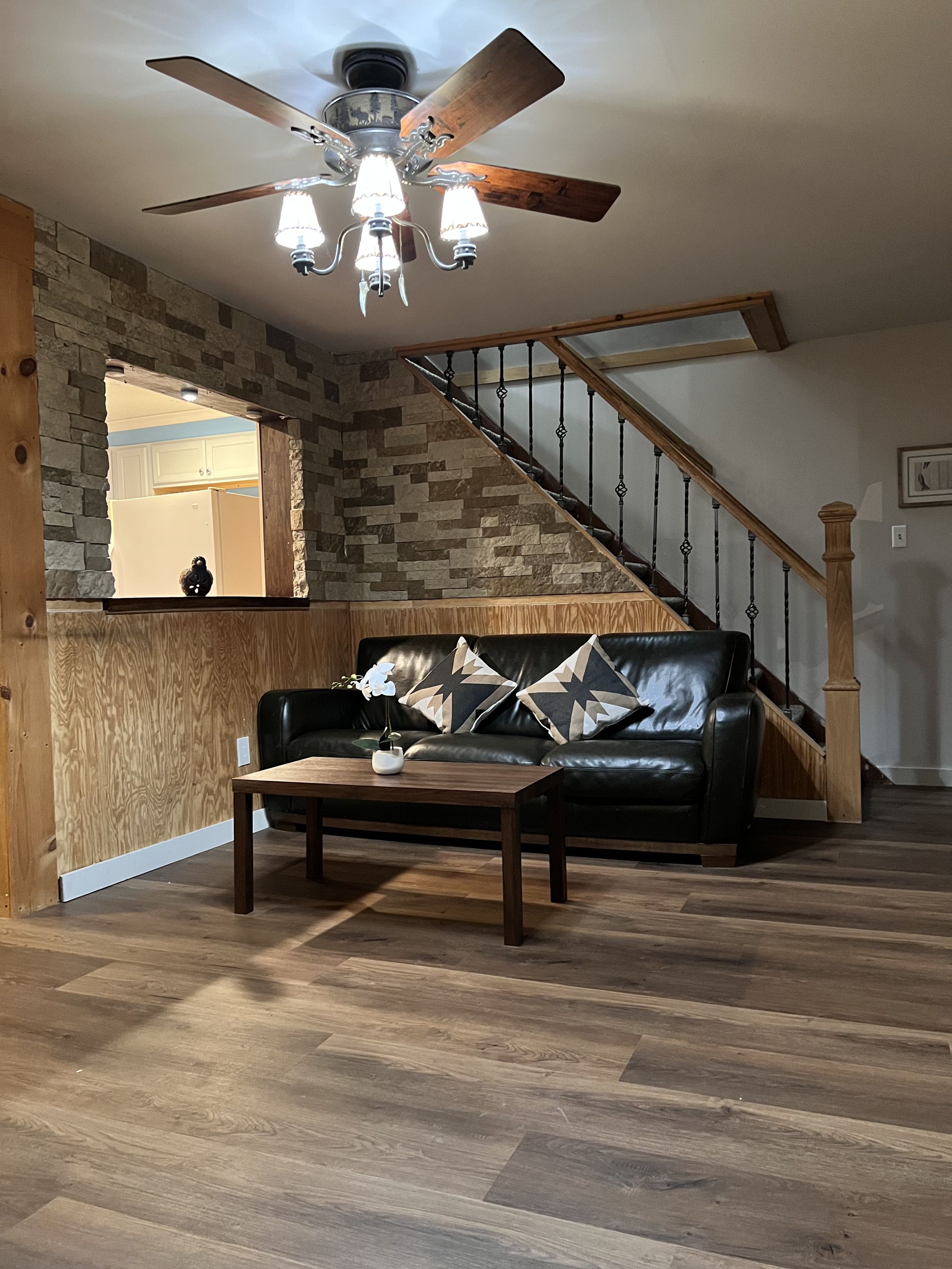 A living room with a wooden floor, a black leather sofa, a wooden coffee table, and a ceiling fan.