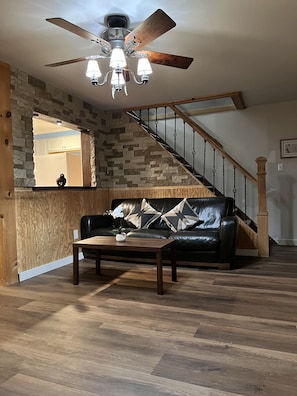 A living room with a wooden floor, a black leather sofa, a wooden coffee table, and a ceiling fan.